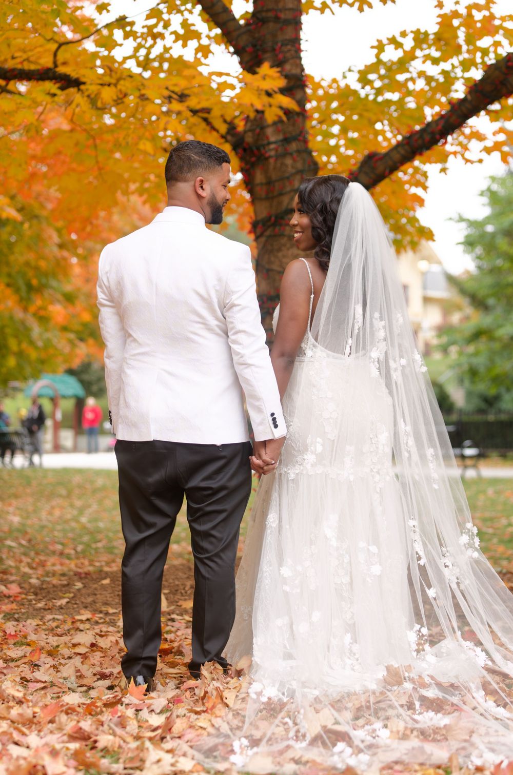 Bride and groom holding hands facing vibrant orange autumn tree, bride in lace gown with long veil, groom in white tux.
