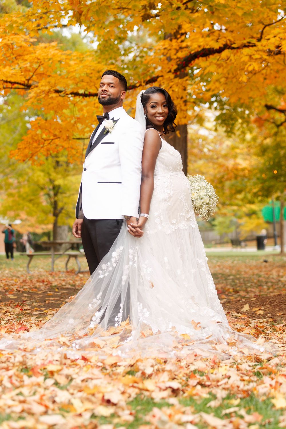 Black bride and groom posing back-to-back in autumn park with golden foliage, bride in lace gown with long floral veil.