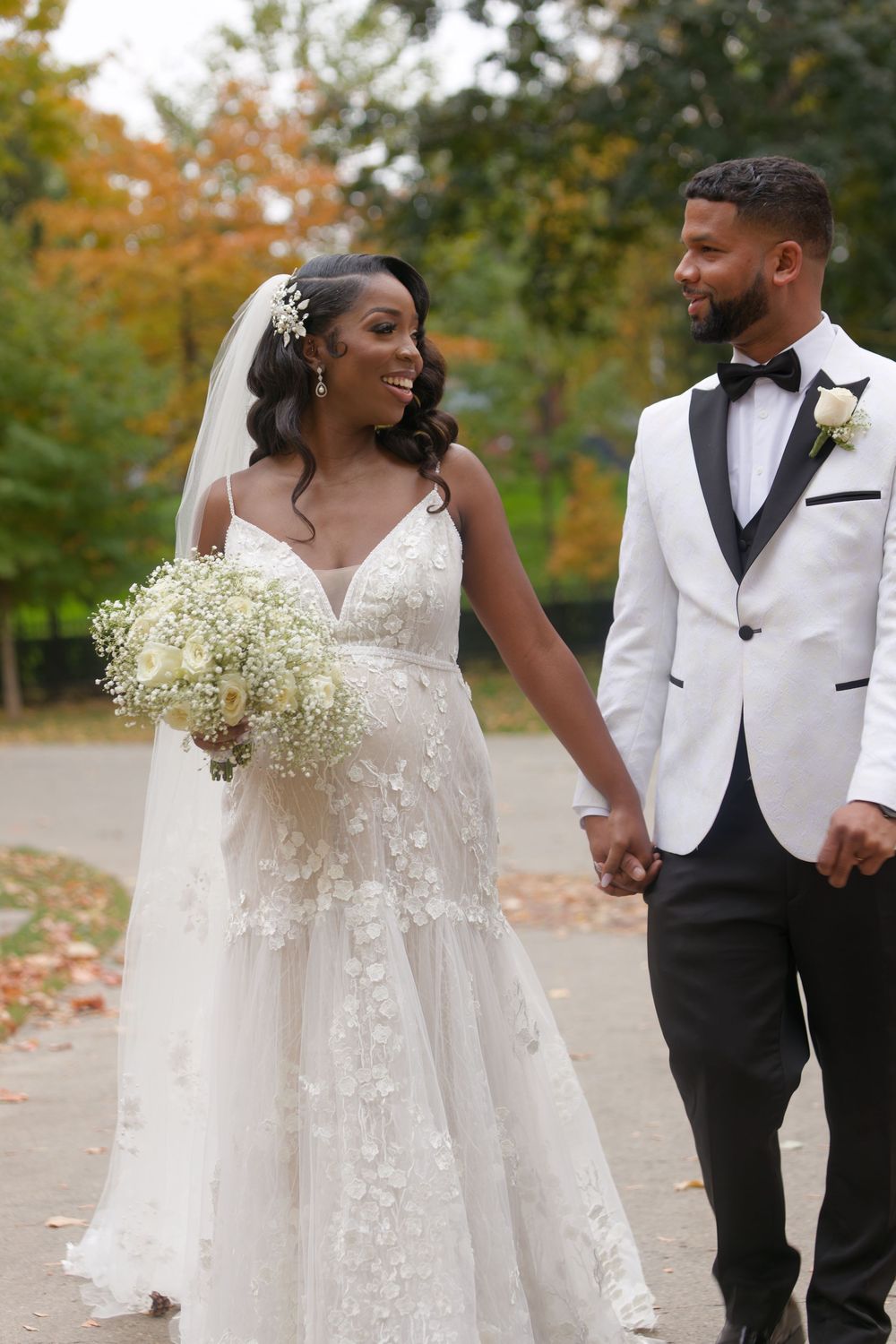 A bride and groom walk hand in hand through a park with vibrant autumn foliage.