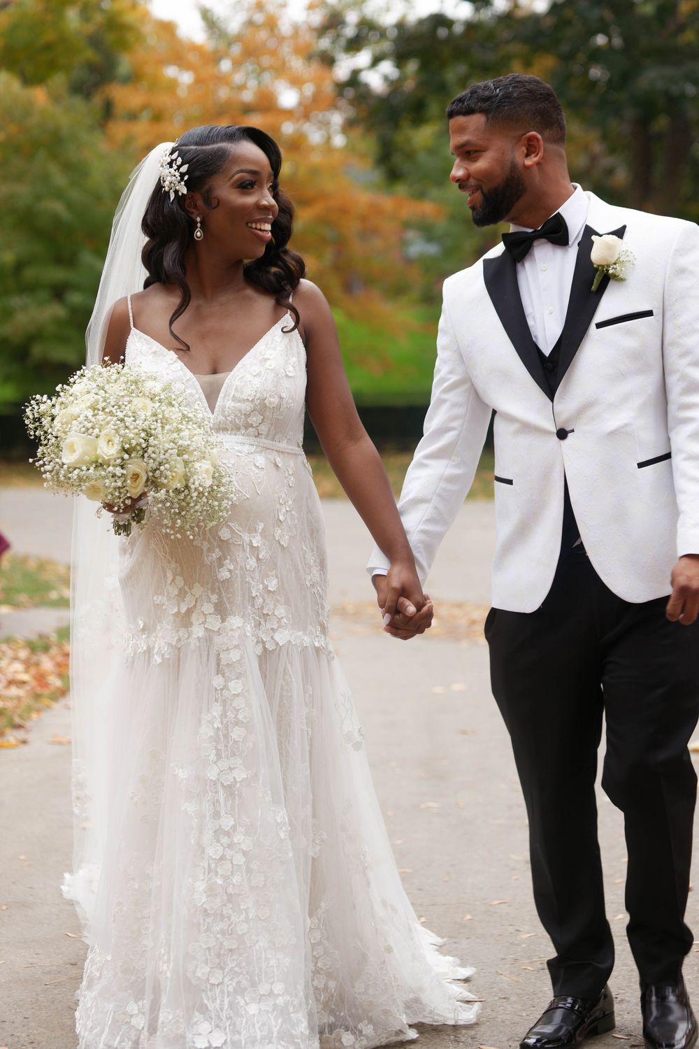 Black bride and groom hold hands walking in autumn park, bride in floral lace gown with bouquet, groom in white tuxedo.
