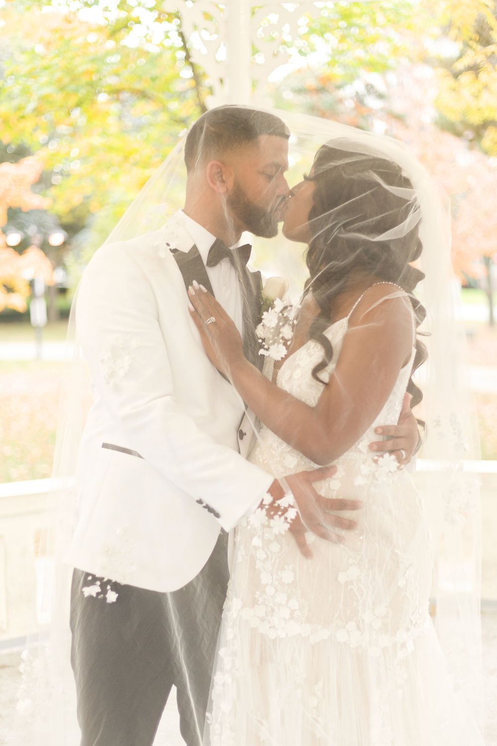 Newlywed couple sharing a kiss under bridal veil at outdoor gazebo, groom in white tuxedo, bride in lace wedding gown.