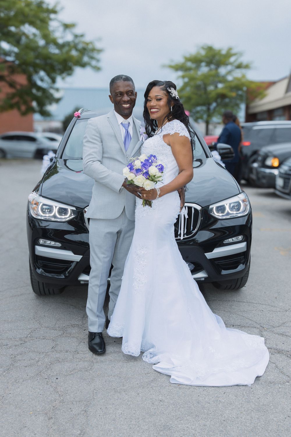 A bride and groom pose proudly in front of a black BMW SUV on their wedding day.