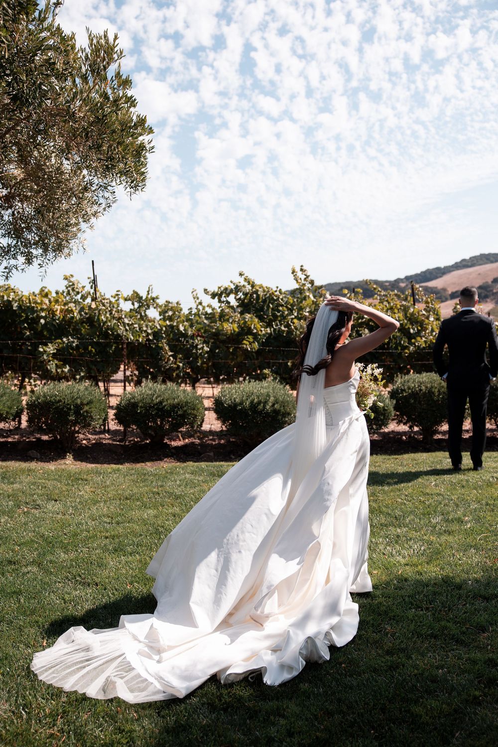 A flowing white wedding dress billows in the breeze at an outdoor vineyard ceremony with rolling hills in the background.
