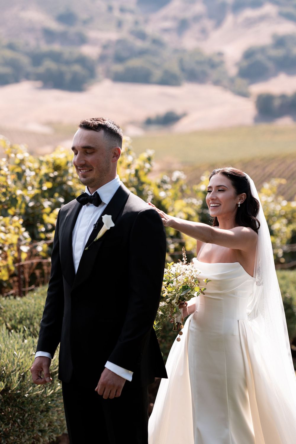 A couple shares joyful moments in their wedding attire against a scenic vineyard backdrop on a sunny afternoon.