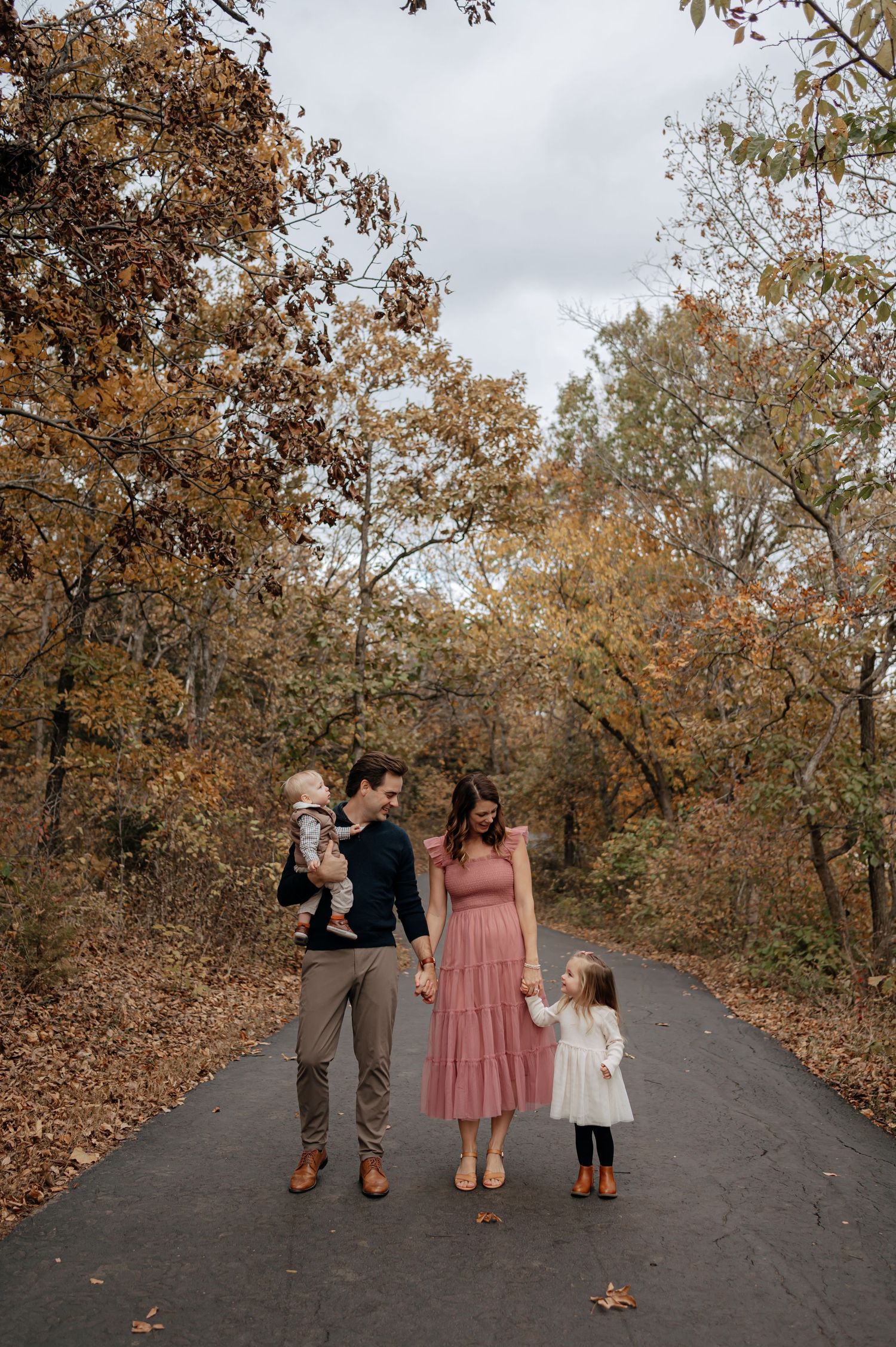 Family of four walking on autumn forest path, parents holding children's hands among fall foliage.