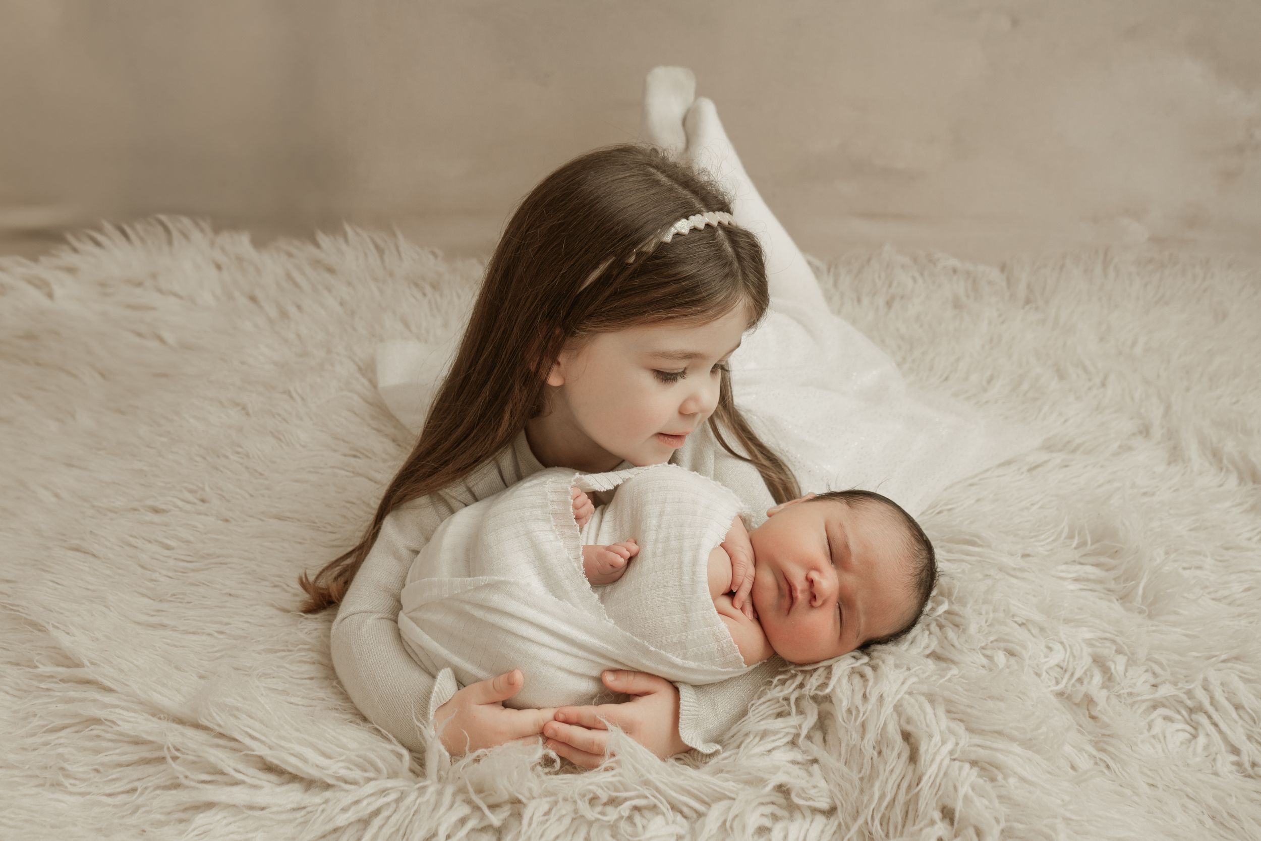 A series of tender moments between a young child and newborn baby on soft white fur backdrop.