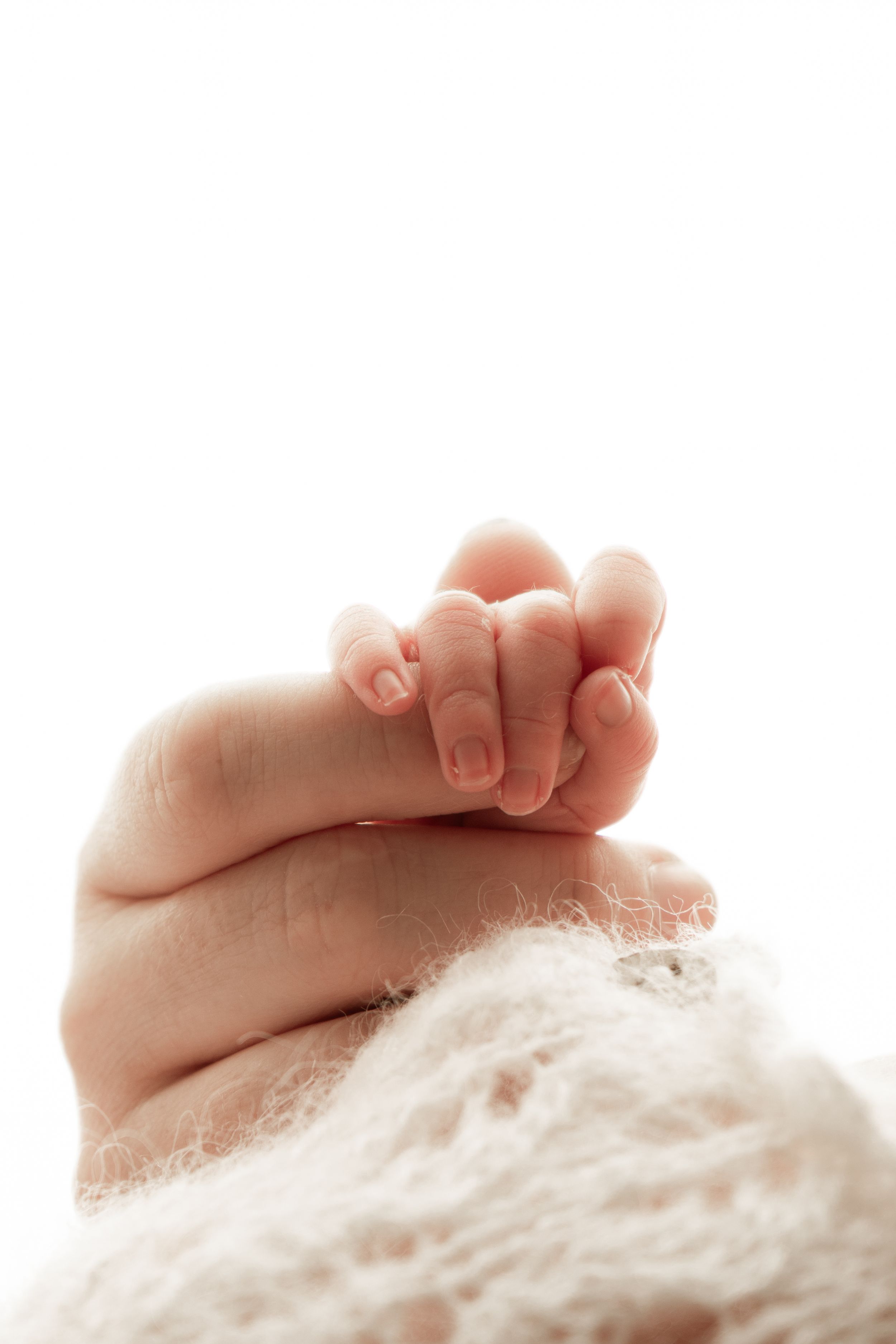 A tiny baby hand gently rests on soft white blanket showing delicate newborn fingers in natural light.
