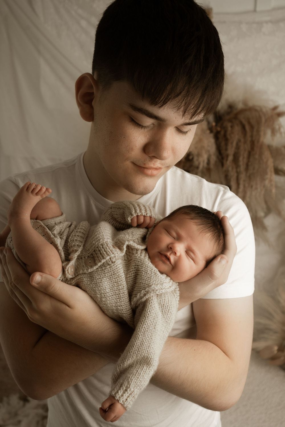 A tender newborn photography session showing a baby wrapped in a soft knit blanket being held against white fabric.