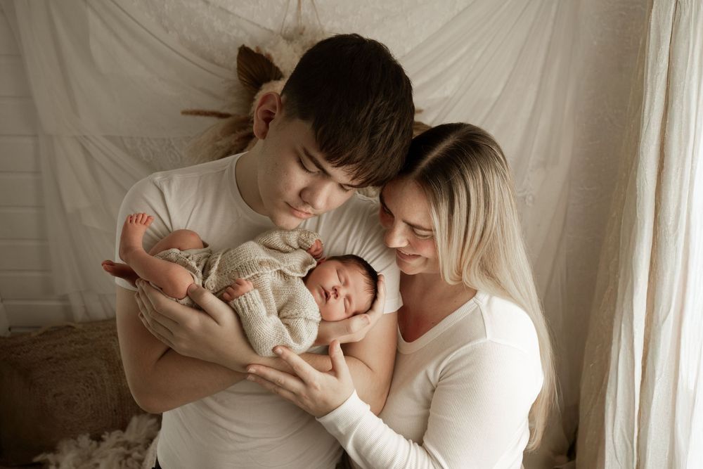 Two parents in white clothing lovingly hold and gaze at their newborn baby against a light fabric backdrop.