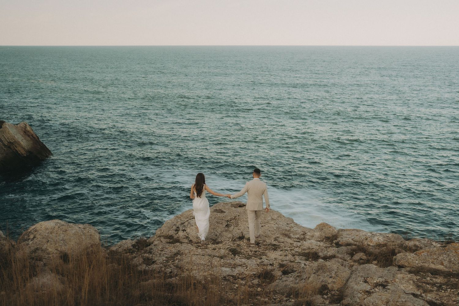 Distant view of couple in white standing on coastal cliff edge overlooking ocean.