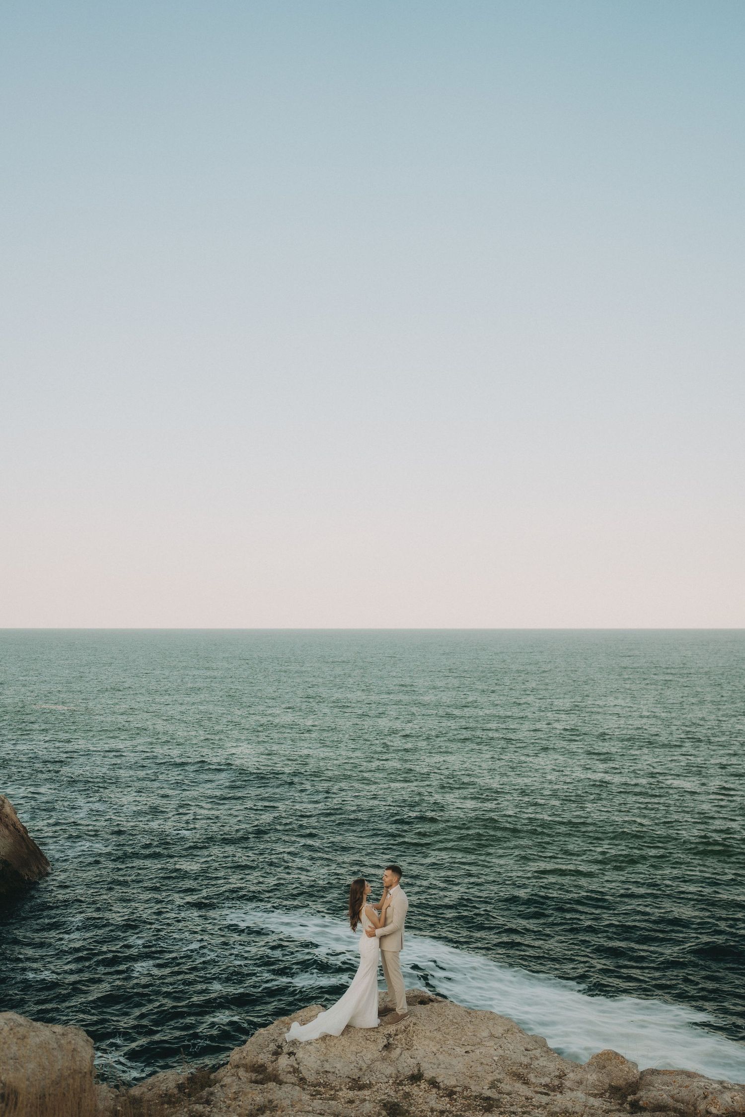 Wide landscape shot of two people in white attire standing on oceanside cliff.