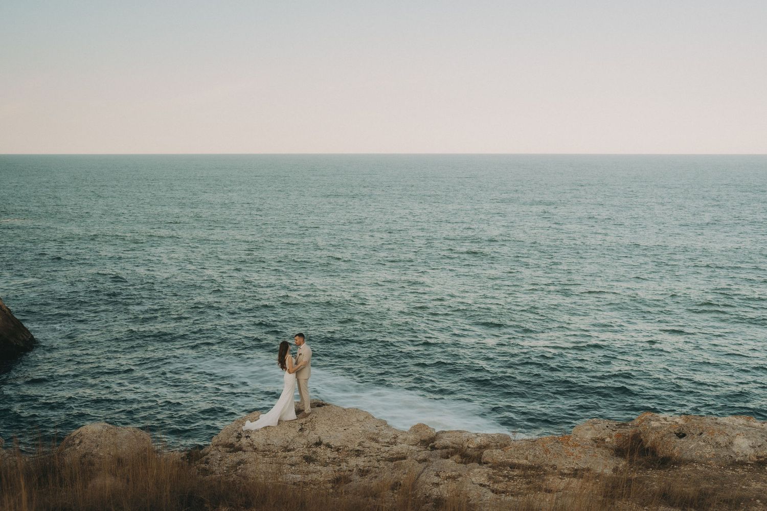 Panoramic coastal scene with figures in white standing on rocky outcrop by ocean.