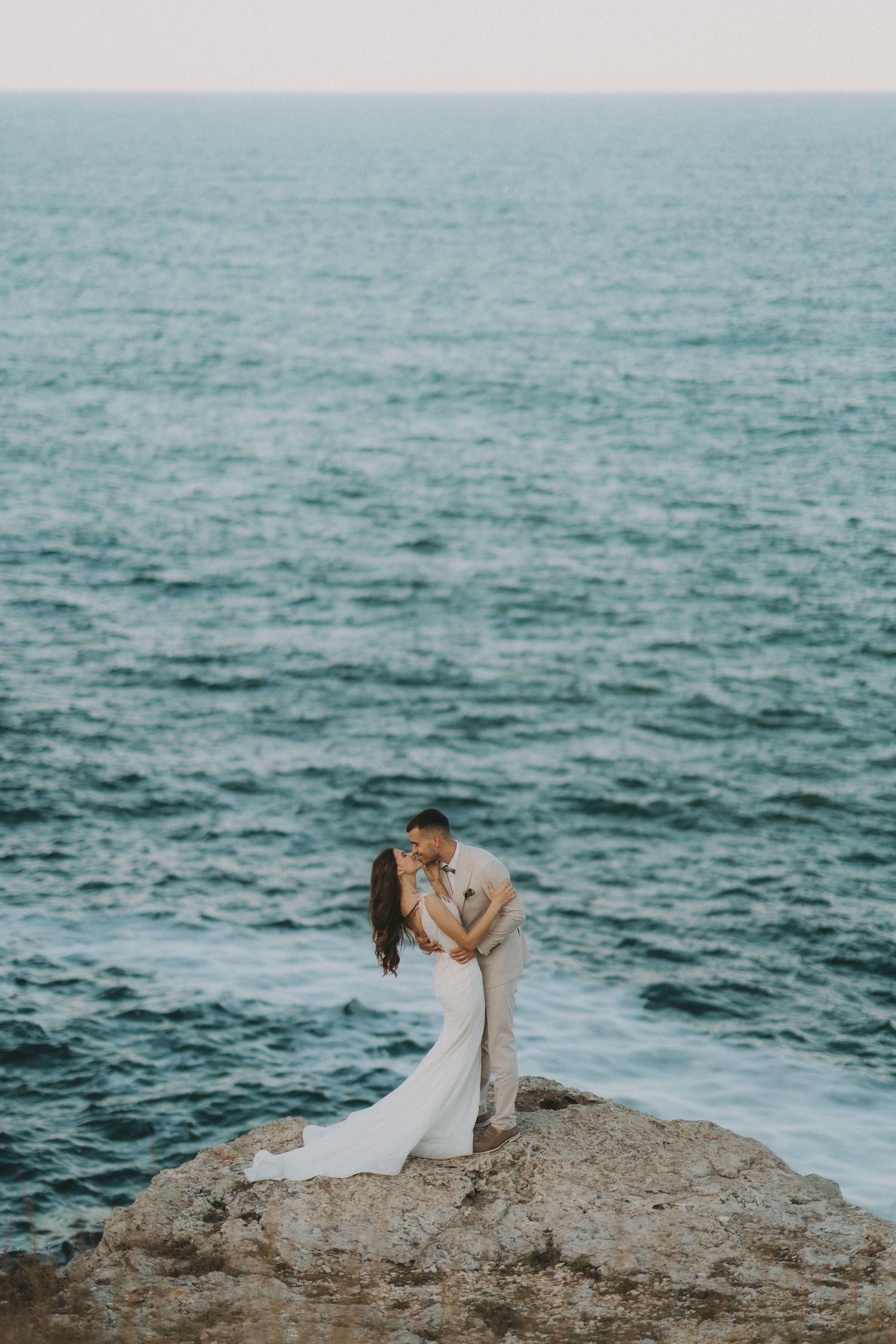 Romantic coastal wedding photo of couple embracing on cliff edge above ocean.