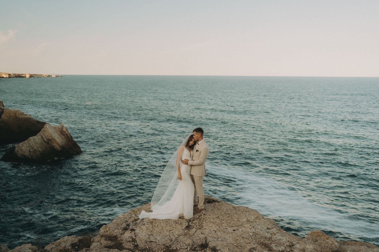 A couple in wedding attire embraces on rocky cliffs overlooking a dramatic ocean vista at sunset.
