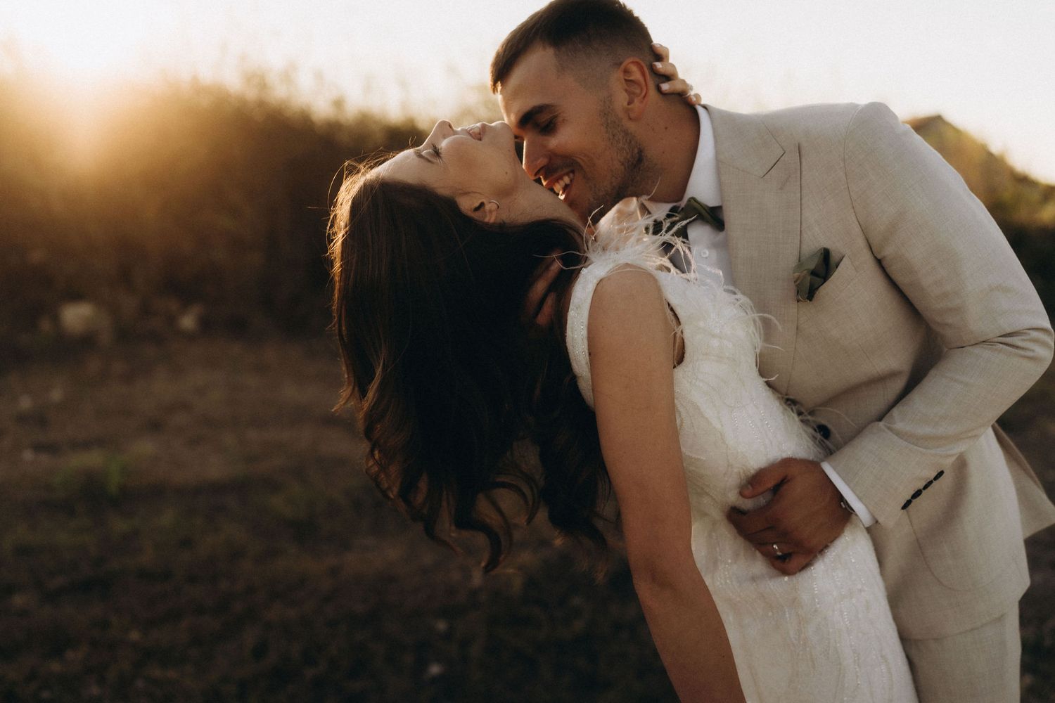 A romantic wedding couple embraces against a golden sunset backdrop near coastal cliffs.