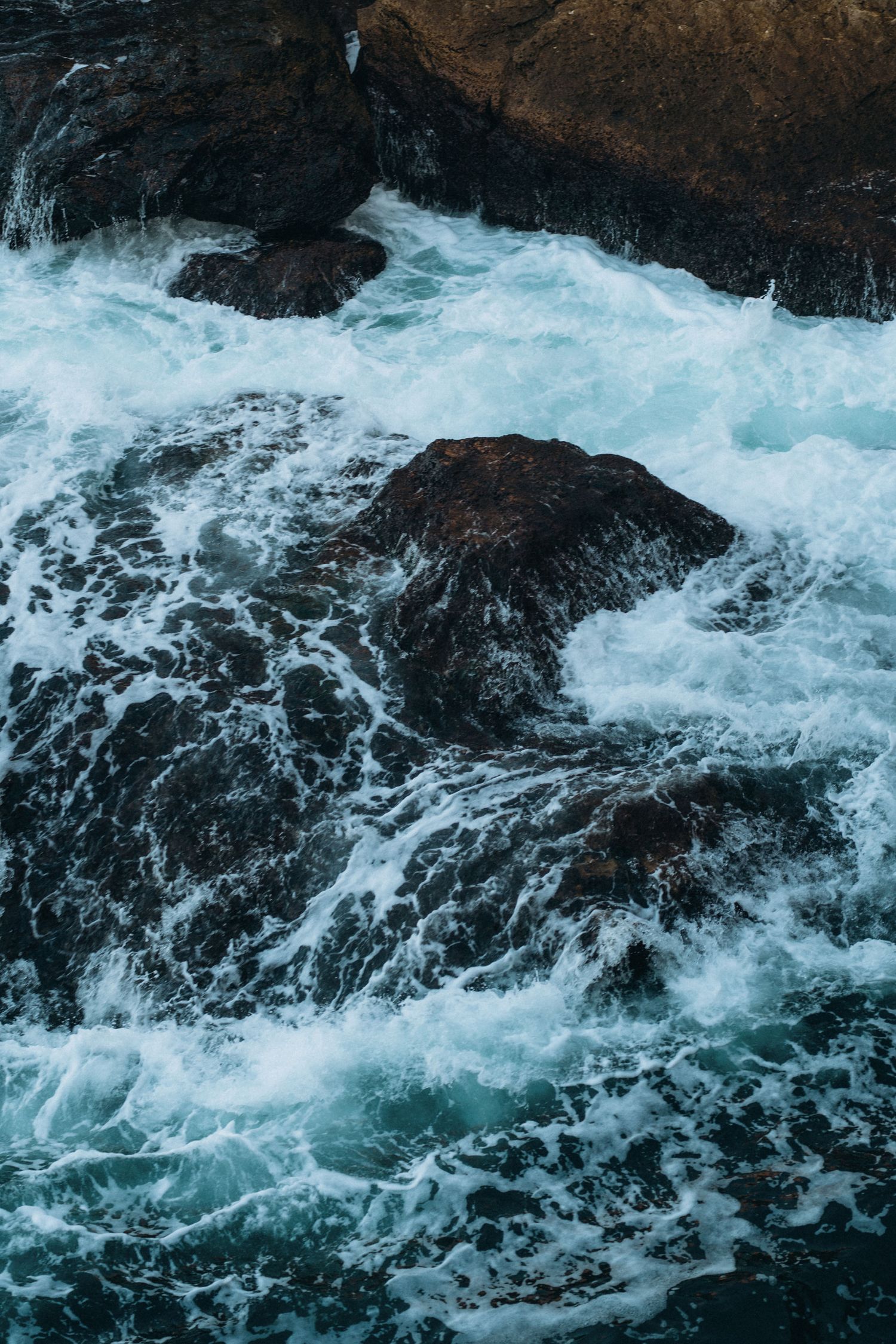 Turquoise waves crash dramatically against dark rocky outcrops in swirling patterns.