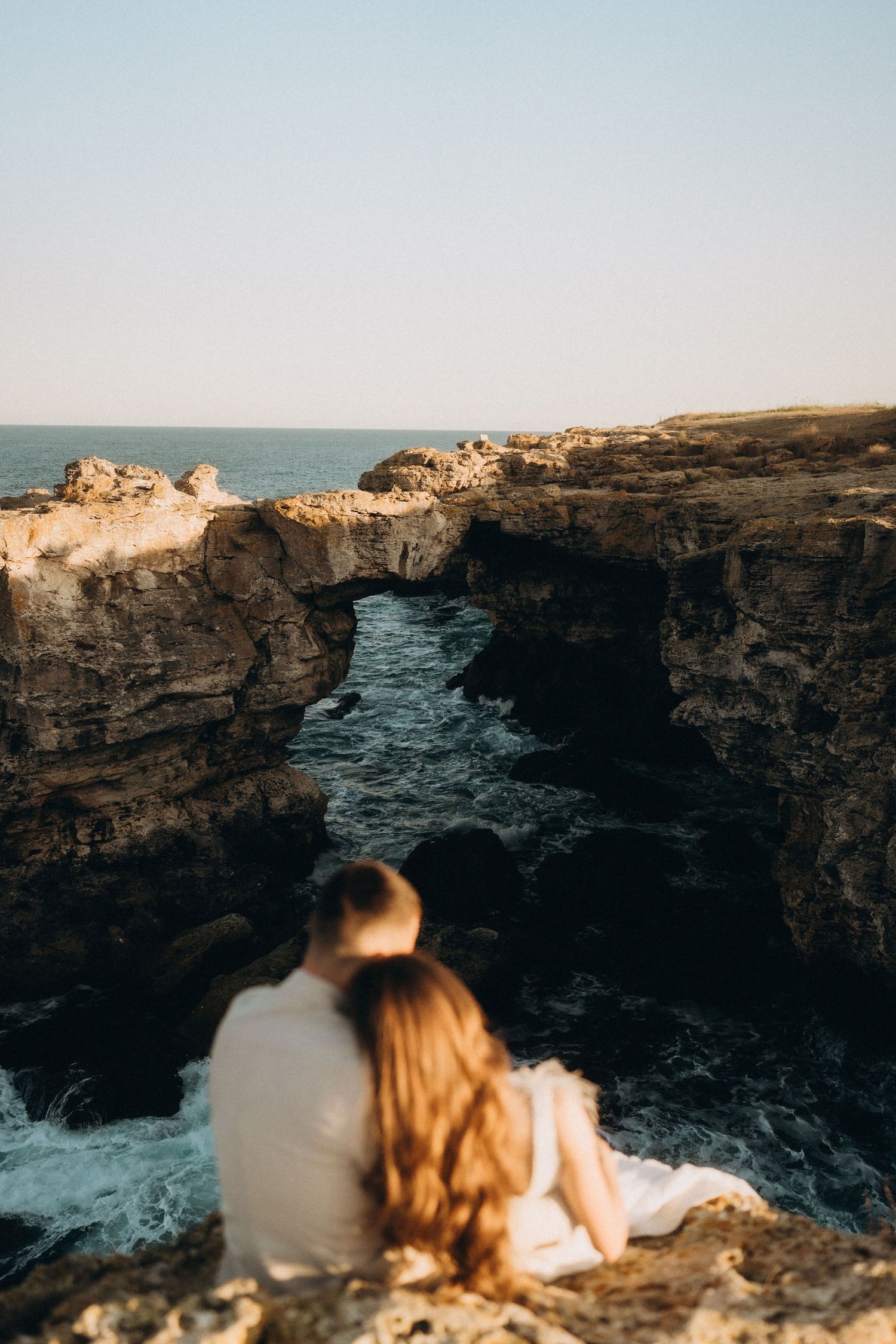A natural stone arch formation over churning ocean waters with a couple enjoying the view at sunset.