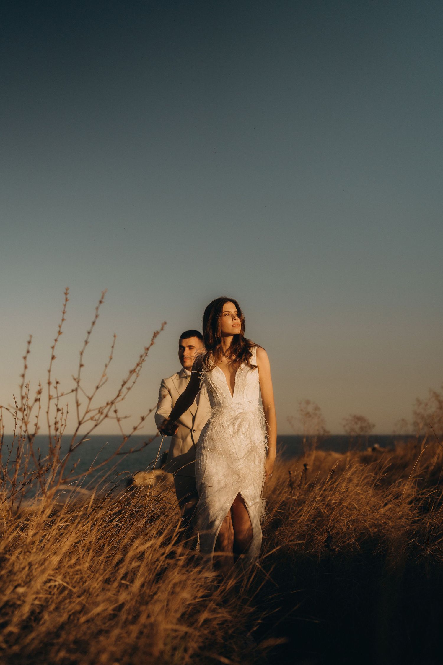 A couple walking through golden grass on a coastal cliff during sunset.