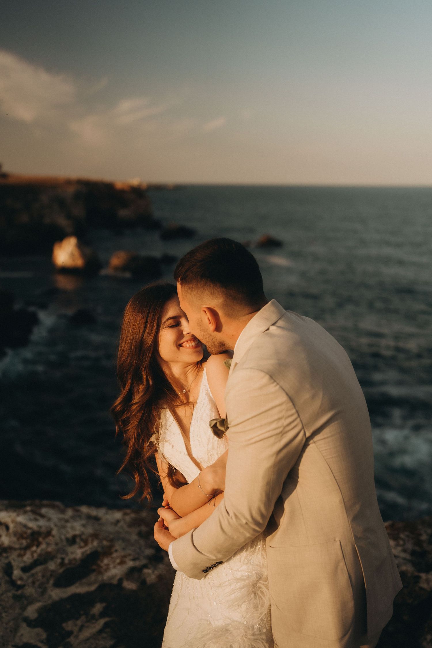 A tender embrace photographed against a dramatic ocean backdrop at sunset.
