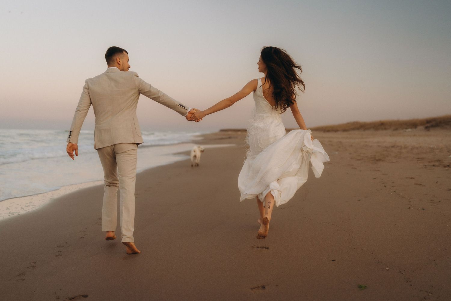 A couple runs playfully hand in hand along the shoreline at dusk.