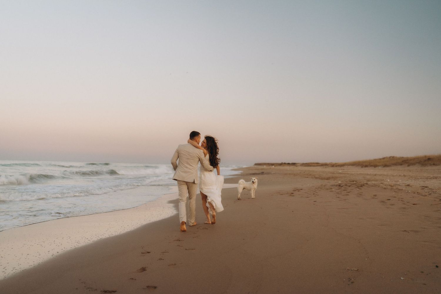 Silhouettes of people walking along a peaceful beach at sunset with a dog in the distance.