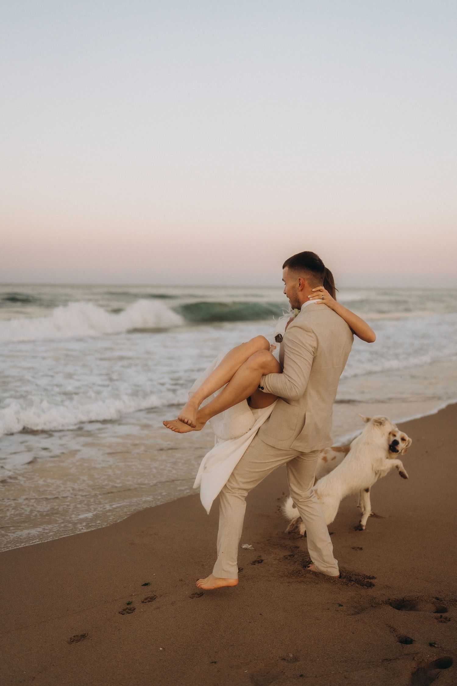 A playful couple dances on the beach with waves and a dog in the background.