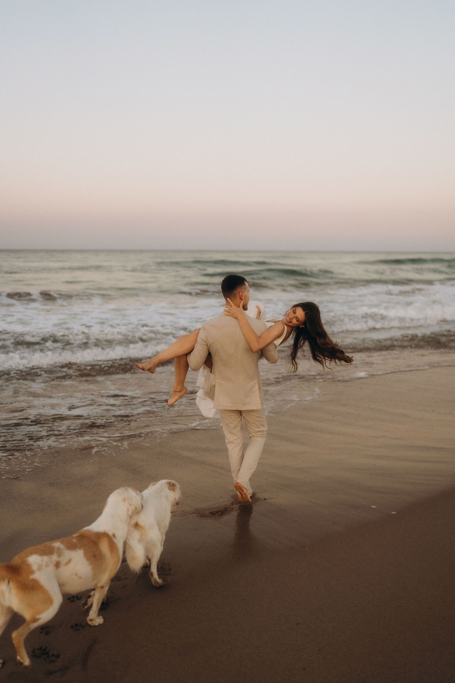 A couple and their dog run playfully along a beach at sunset with waves crashing in the background.