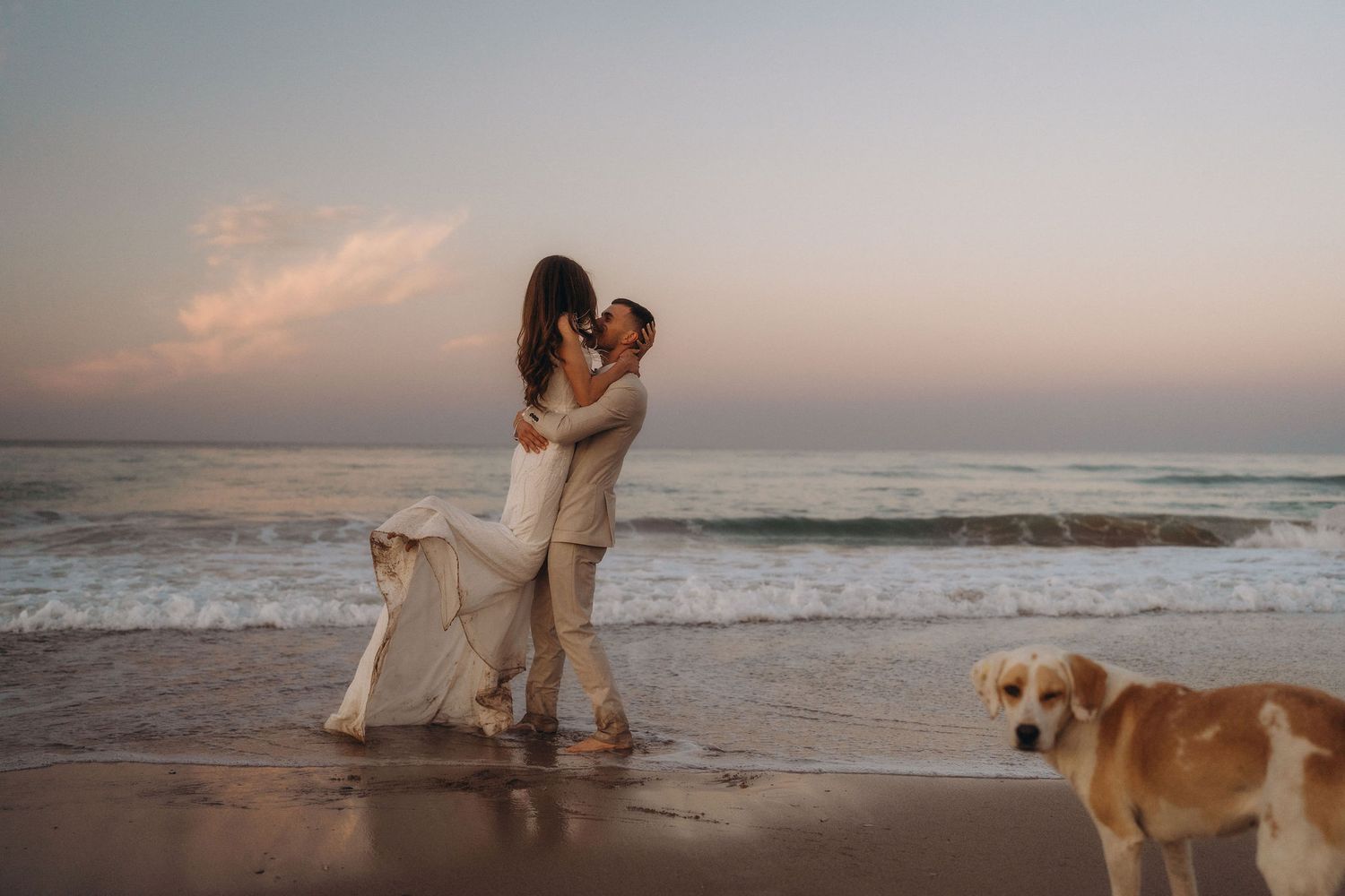 A couple embraces on the shoreline while their dog watches during golden hour.