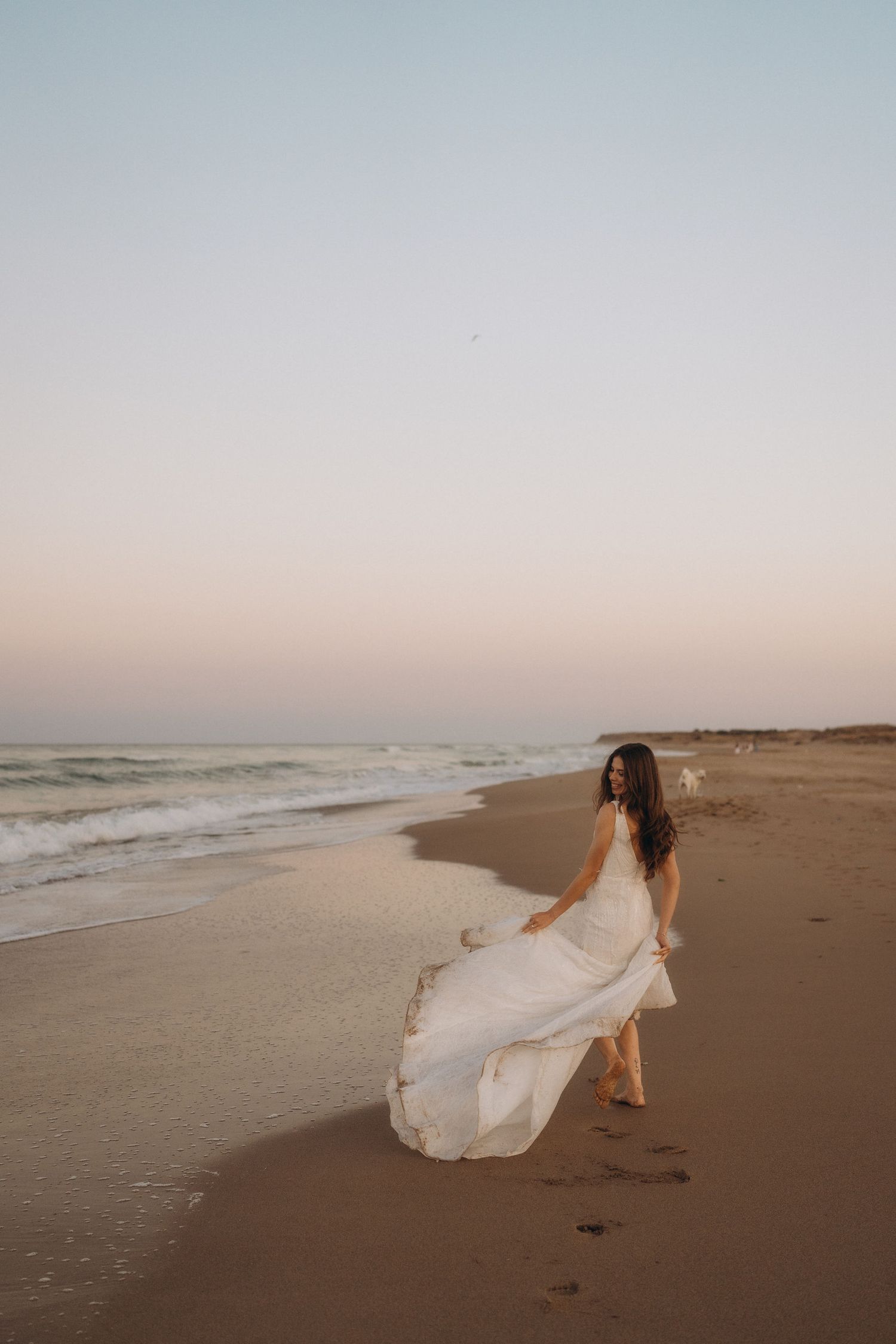 A flowing white dress catches the wind as a couple walks along an empty beach at sunset.