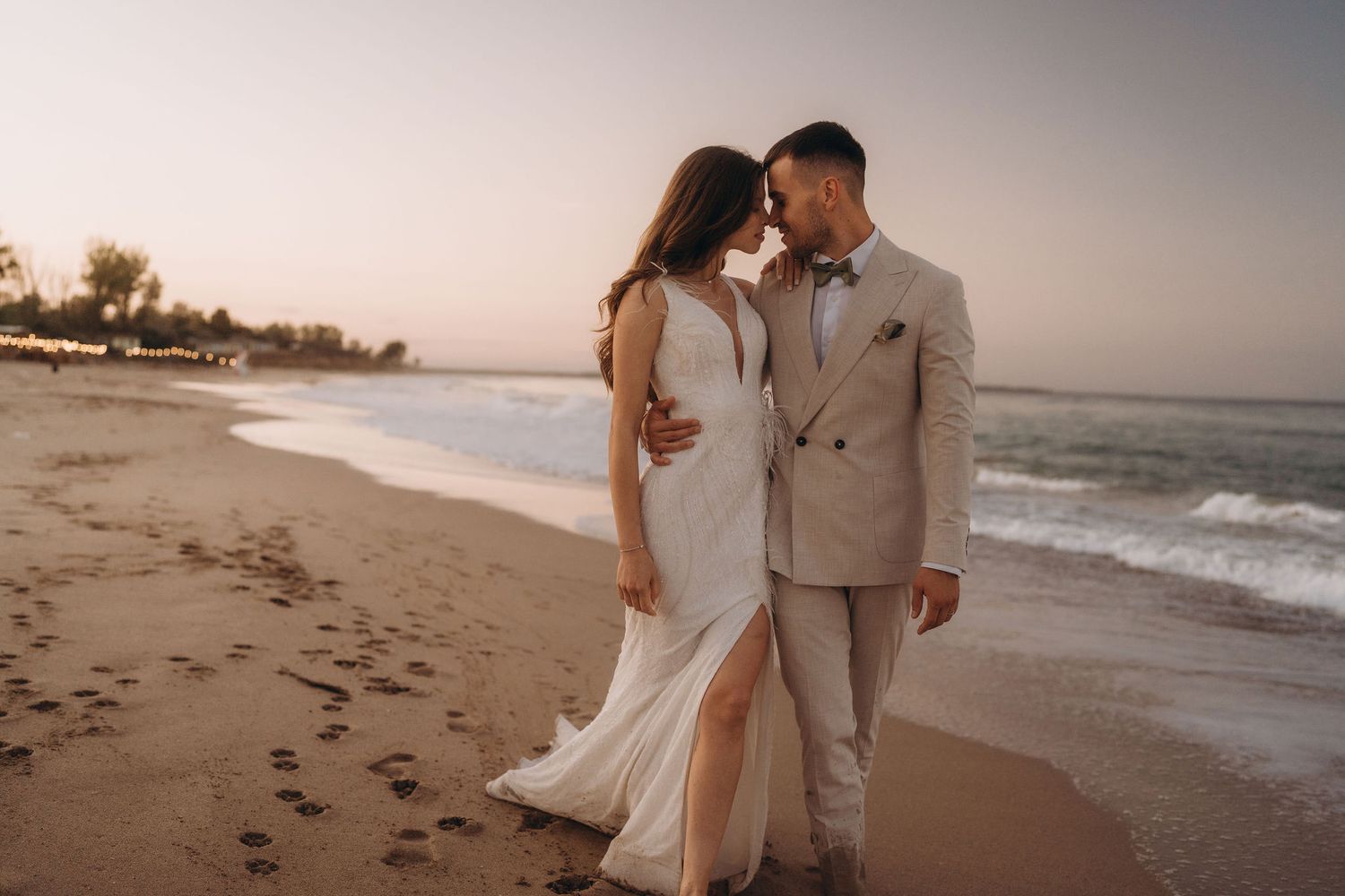 A couple in light-colored attire walks along the beach shoreline during golden hour.
