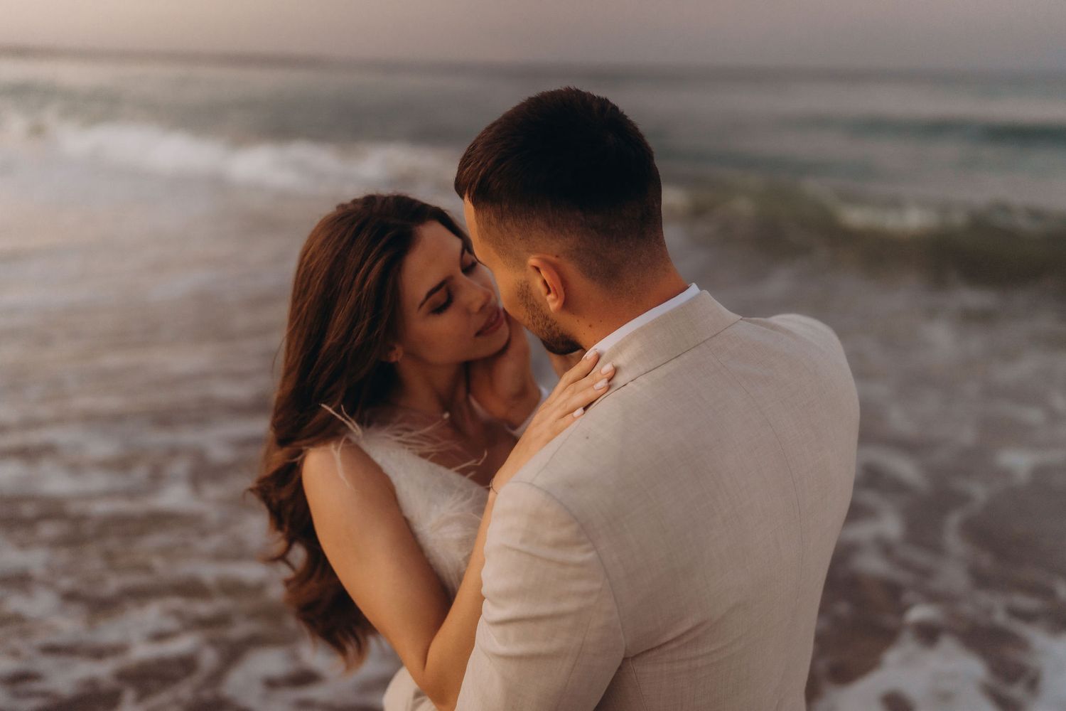 A romantic couple shares an intimate moment on a beach during sunset in neutral-toned attire.
