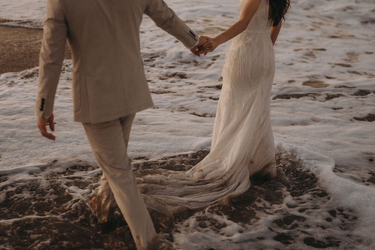 A couple walks hand in hand along the shoreline wearing beige and white outfits.