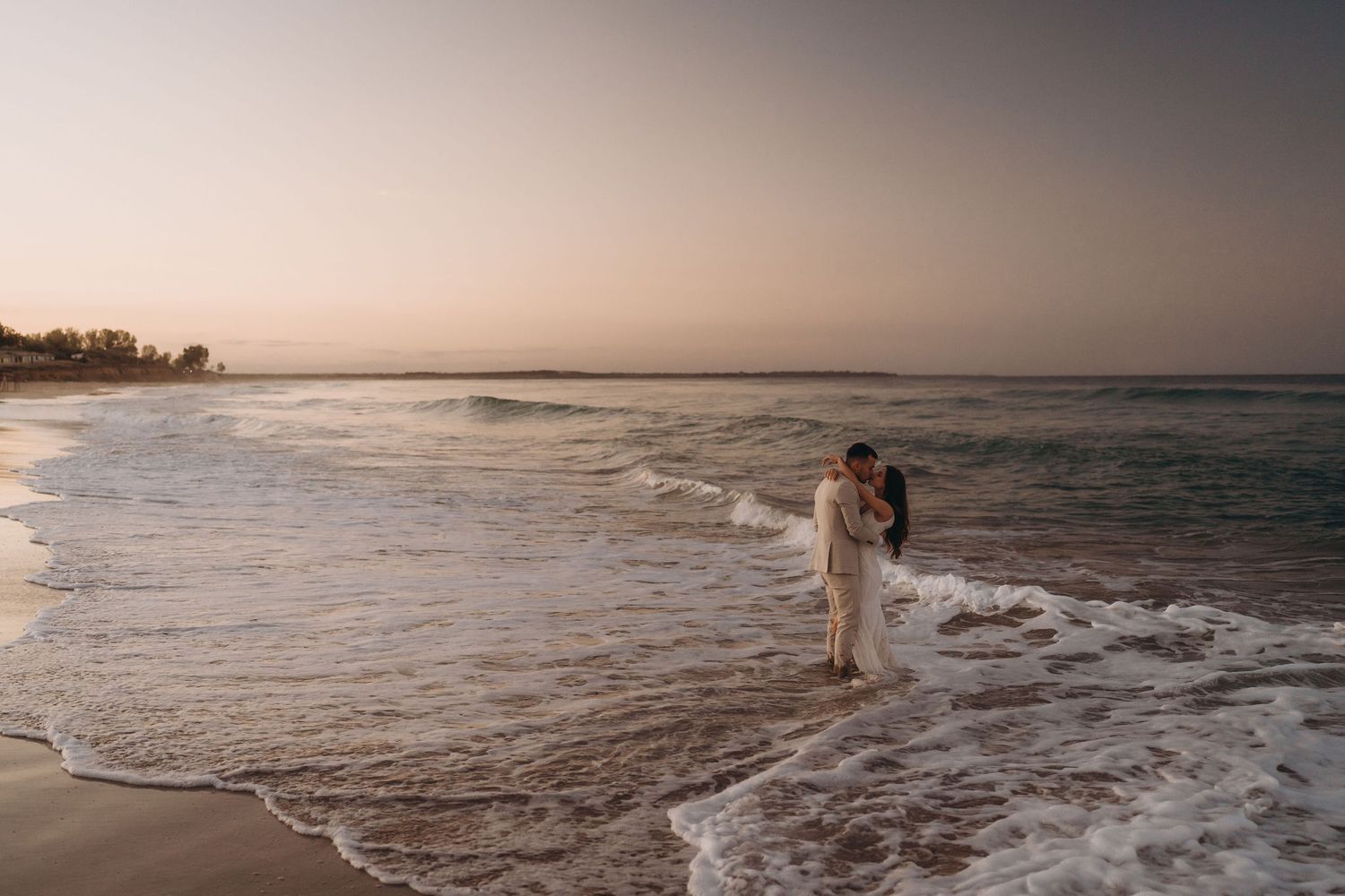 A couple stands in shallow ocean waves at sunset creating a romantic beach scene.