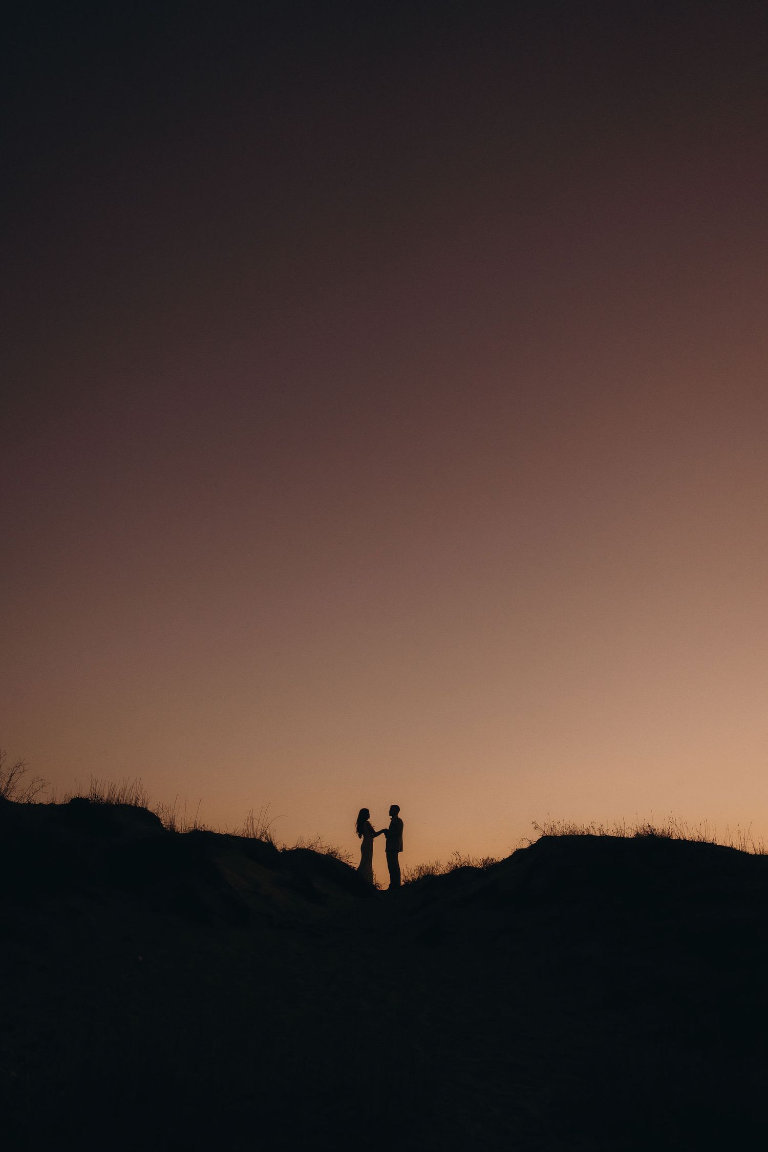 Two silhouettes stand on hilltop against dramatic sunset sky.