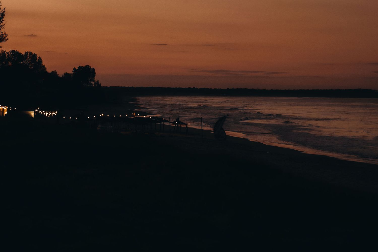 Coastal silhouette at sunset with distant city lights twinkling along the shoreline.