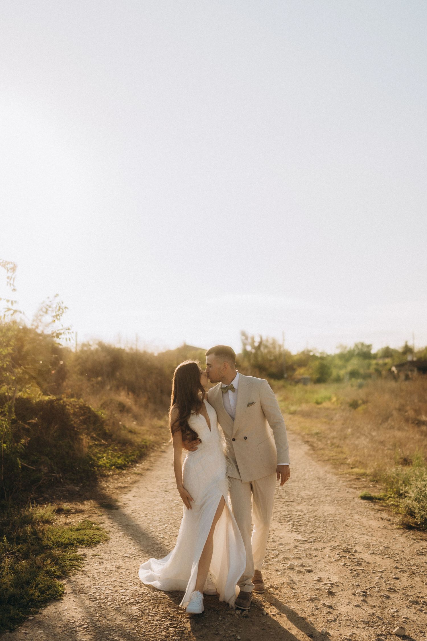 A couple in wedding attire walks along a sunlit dirt path surrounded by natural foliage.
