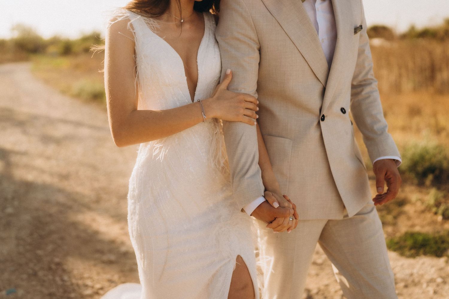 Couple in light-colored wedding attire walking hand in hand on a rural path.