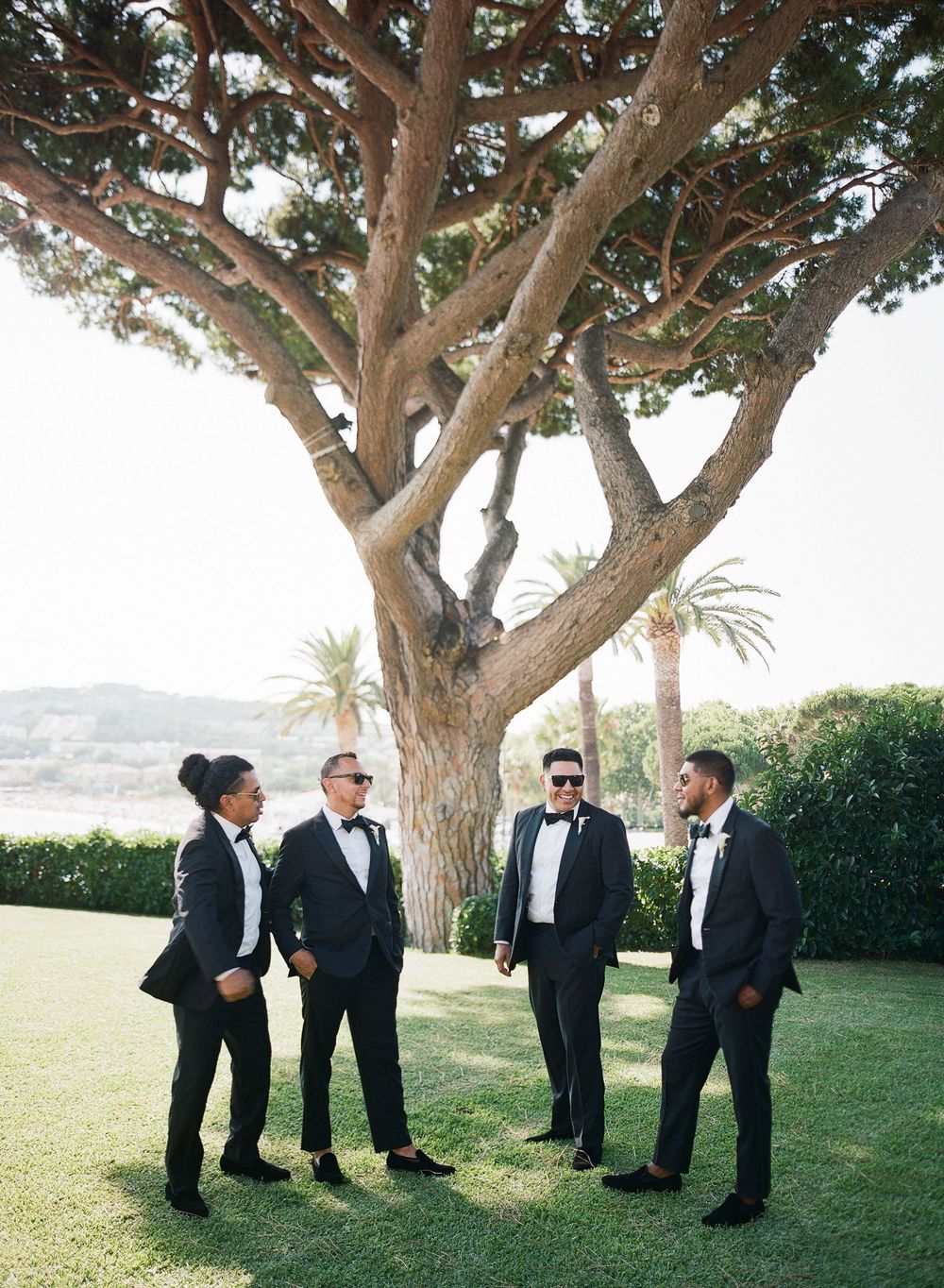 A group of men in black tuxedos gather beneath large trees on a grassy hillside for a formal outdoor event.