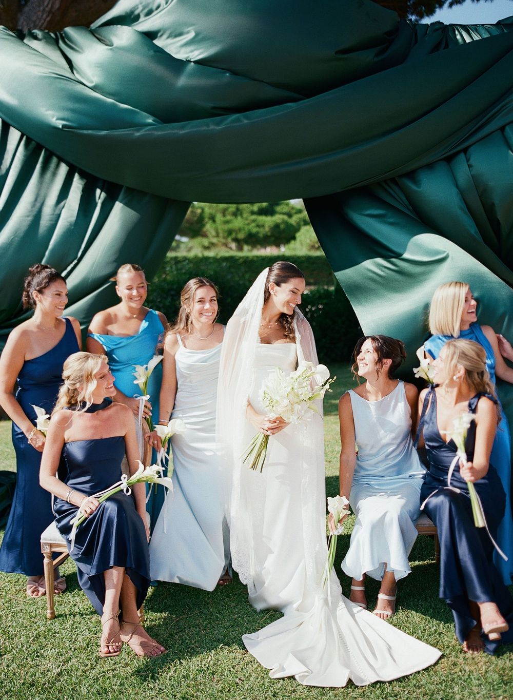 Wedding party in blue and white dresses laughing together while holding a large teal parachute outdoors.