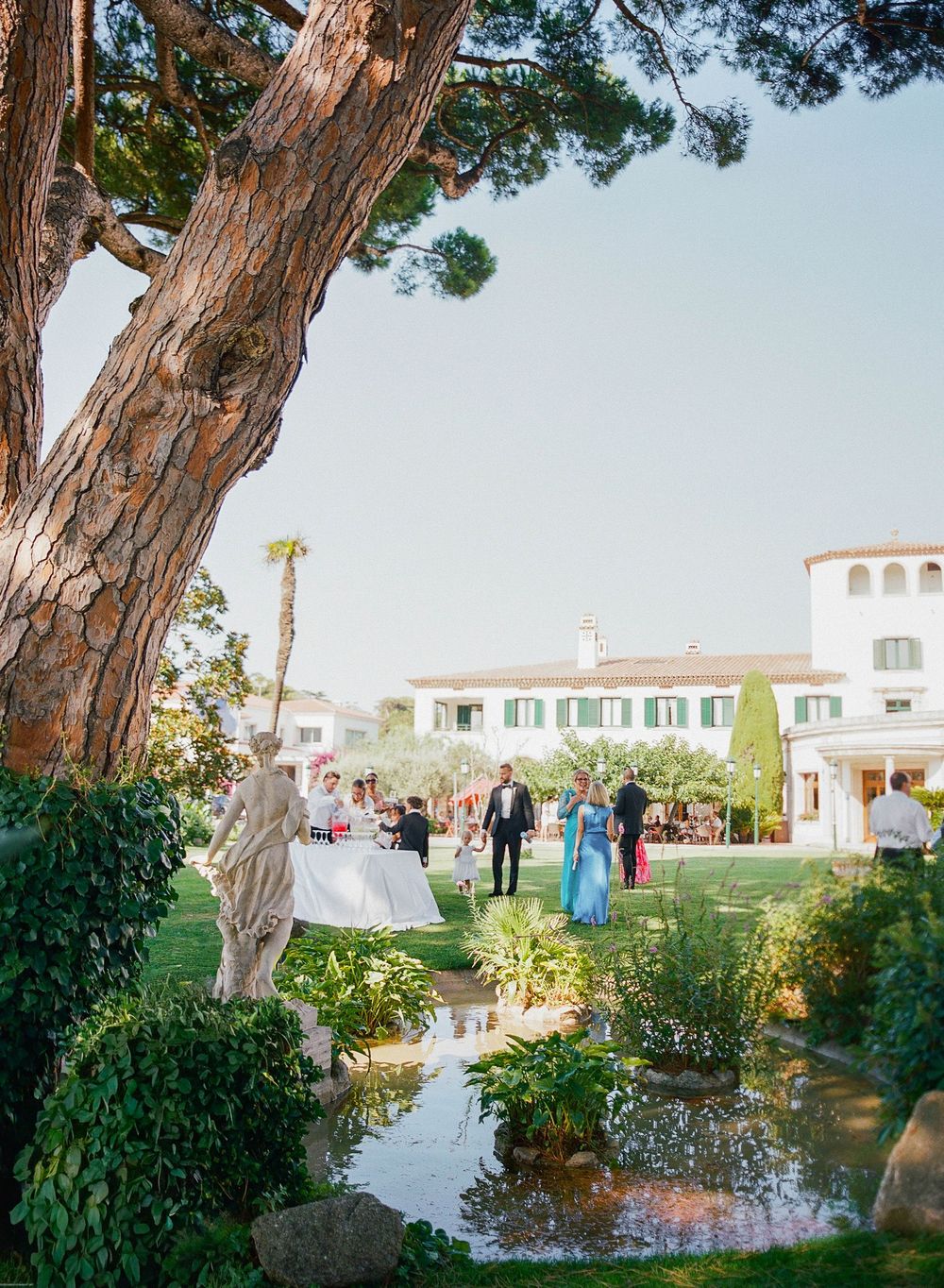 Wedding guests gather on a scenic terrace with tall cypress trees and Mediterranean-style architecture in the background.