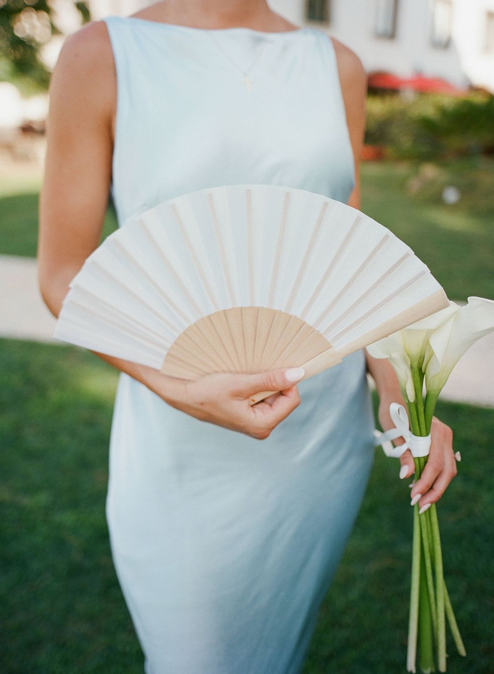 Someone in a light blue dress holds white calla lilies and a delicate folding fan outdoors.