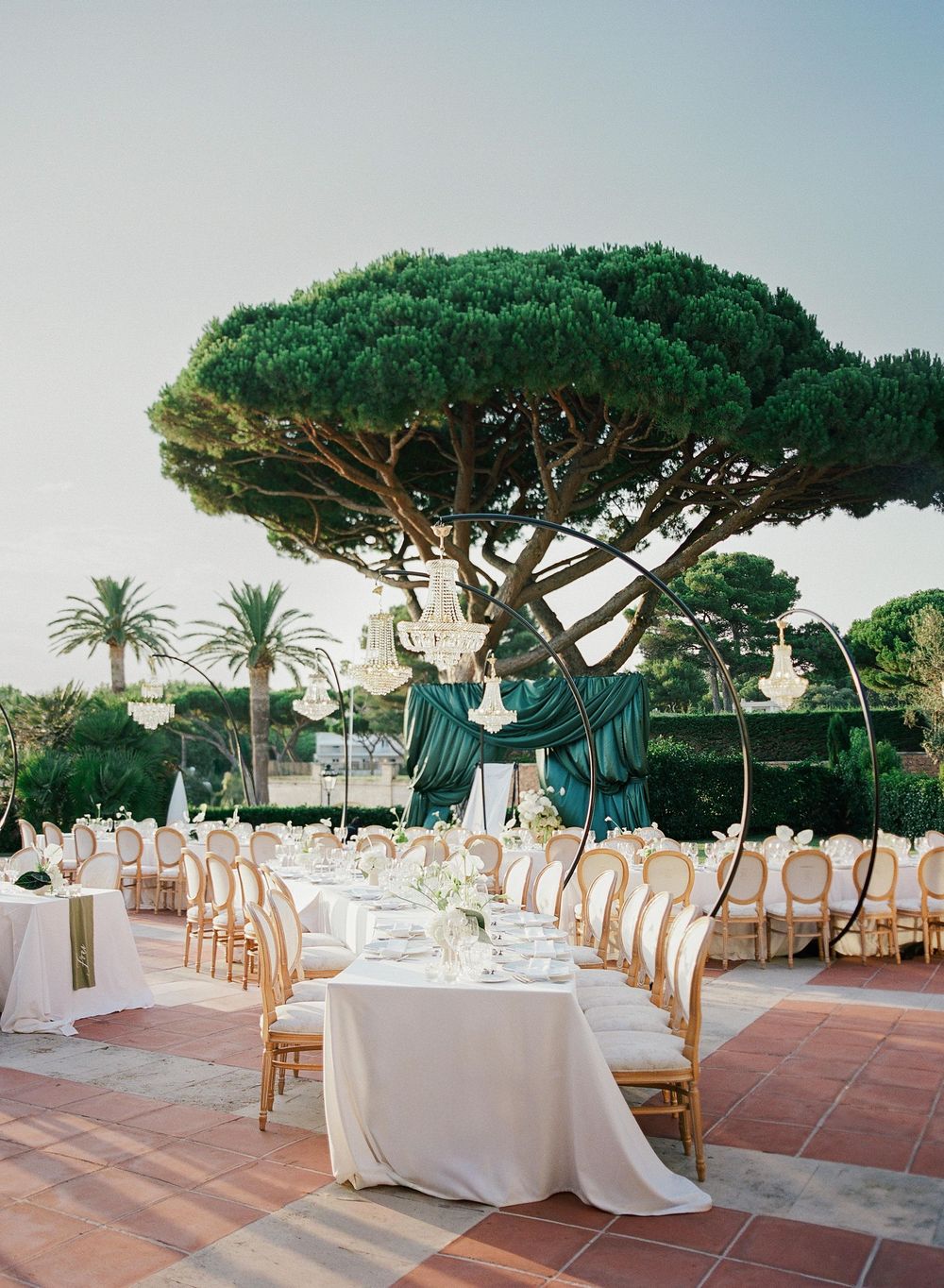 Elegant outdoor wedding reception setup with white-clothed tables and wooden chairs under Mediterranean pine trees.