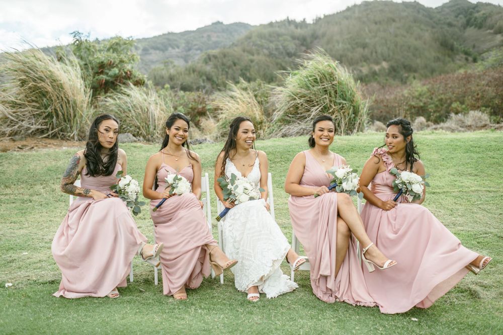 Bridesmaids in blush pink dresses pose playfully on grassy hillside with mountain backdrop.