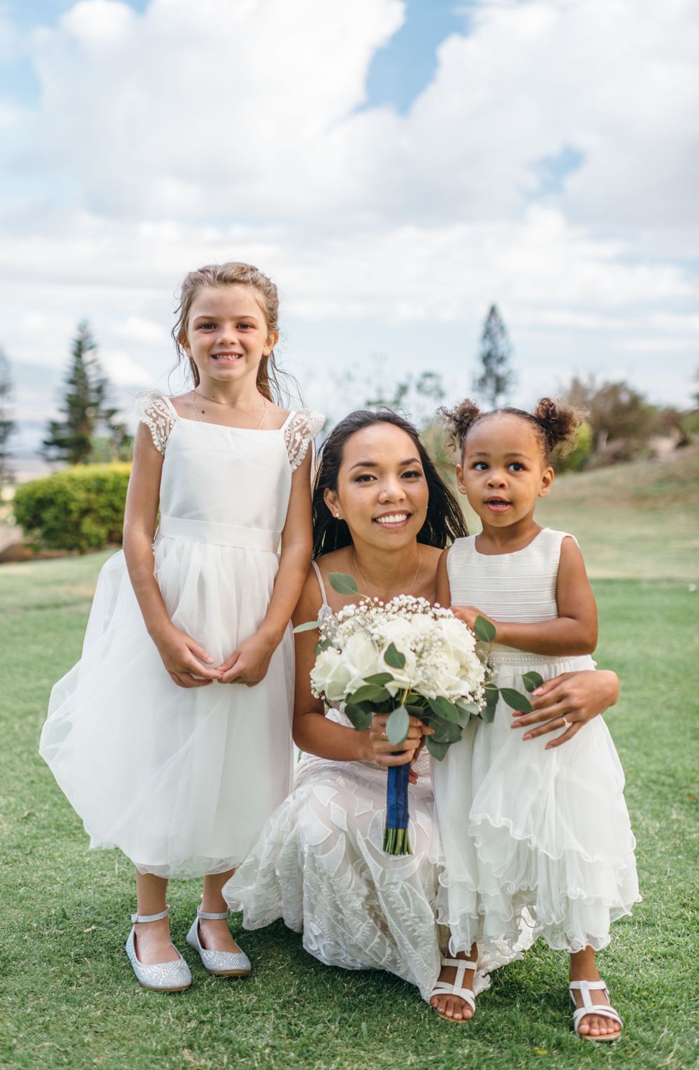 Young flower girls in white dresses pose with bouquets on sunny lawn.