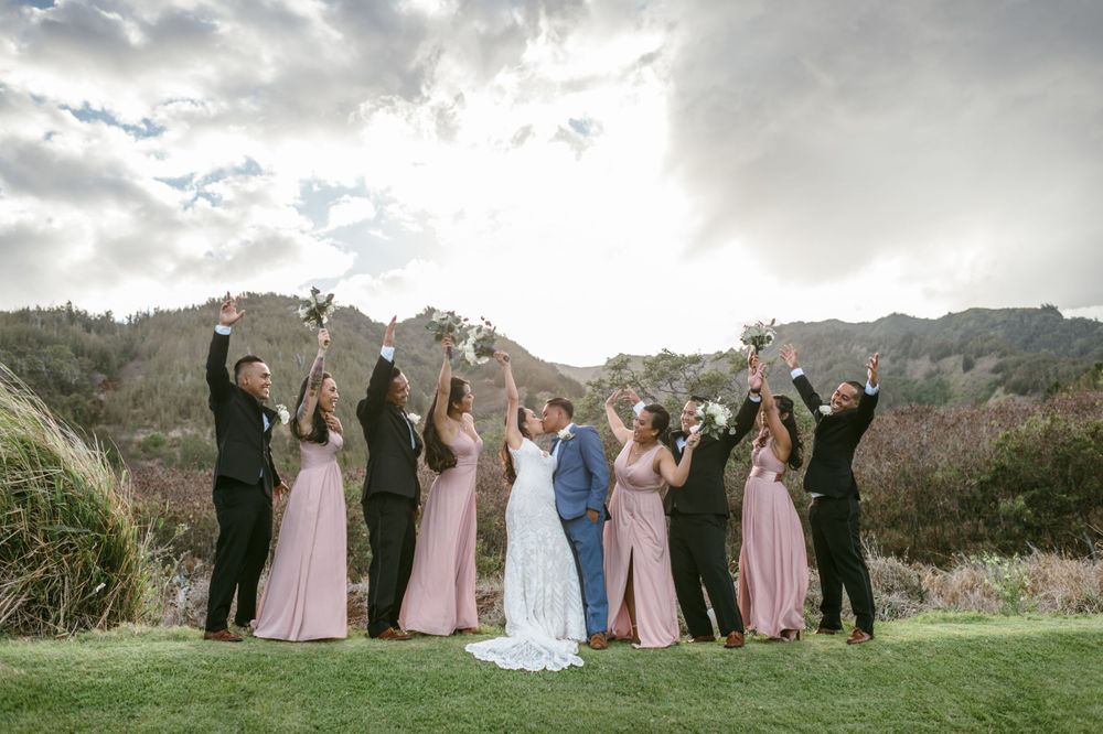 Wedding party celebrates with arms raised against mountainous backdrop at outdoor venue.