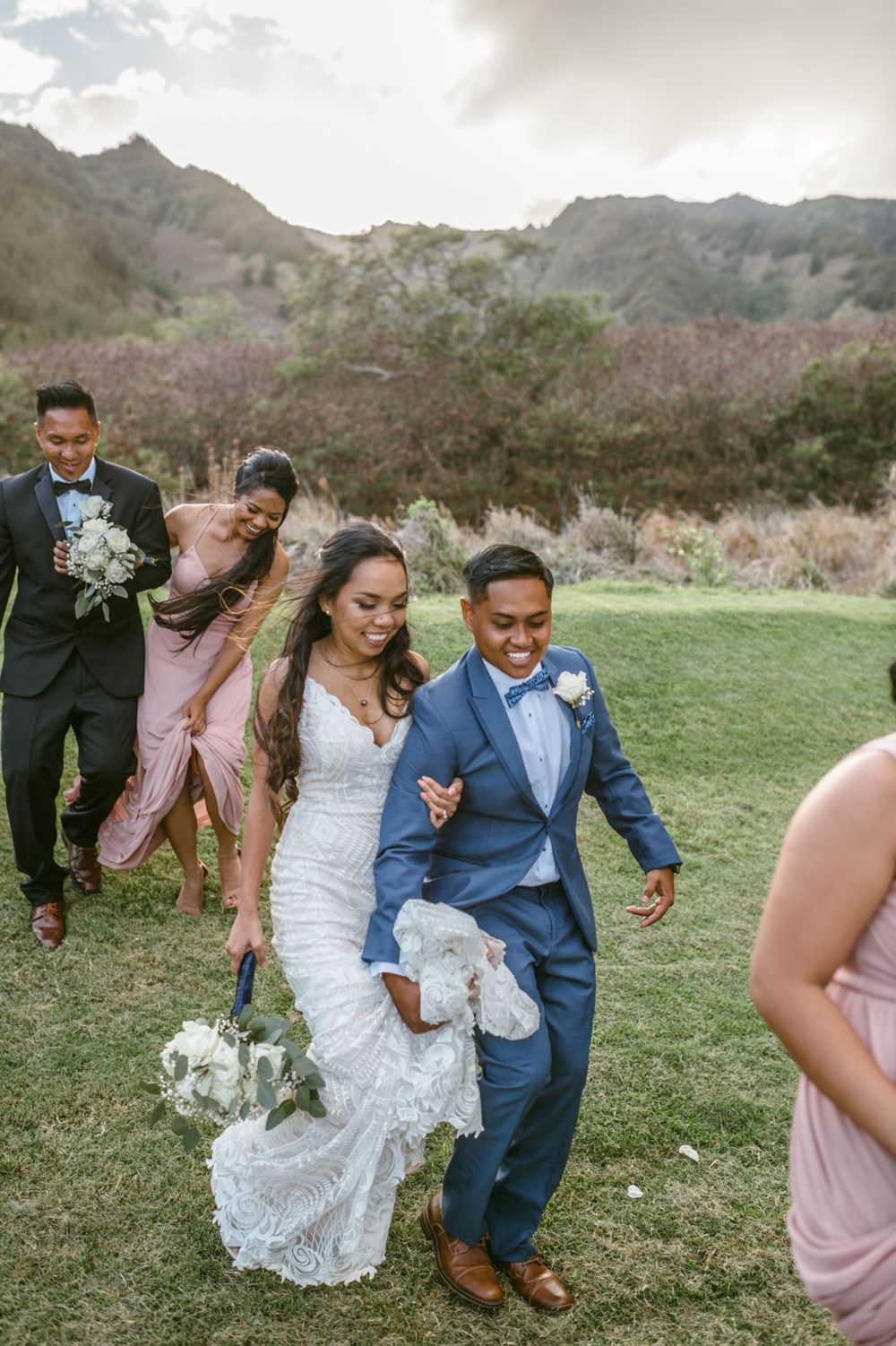 Wedding party walks across grass with mountains in background during outdoor celebration.