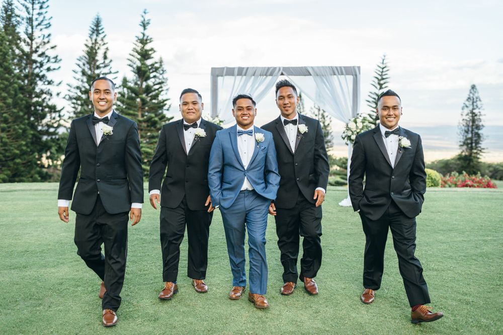 Five groomsmen in formal attire pose together on grass with evergreen trees in background.