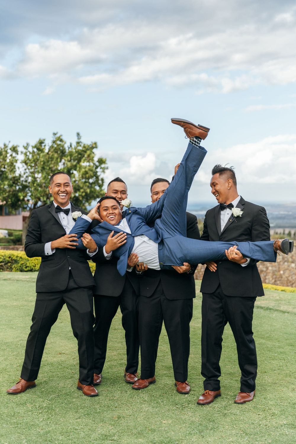 Groomsmen in black tuxedos playfully lift and hold one of their group members horizontally.