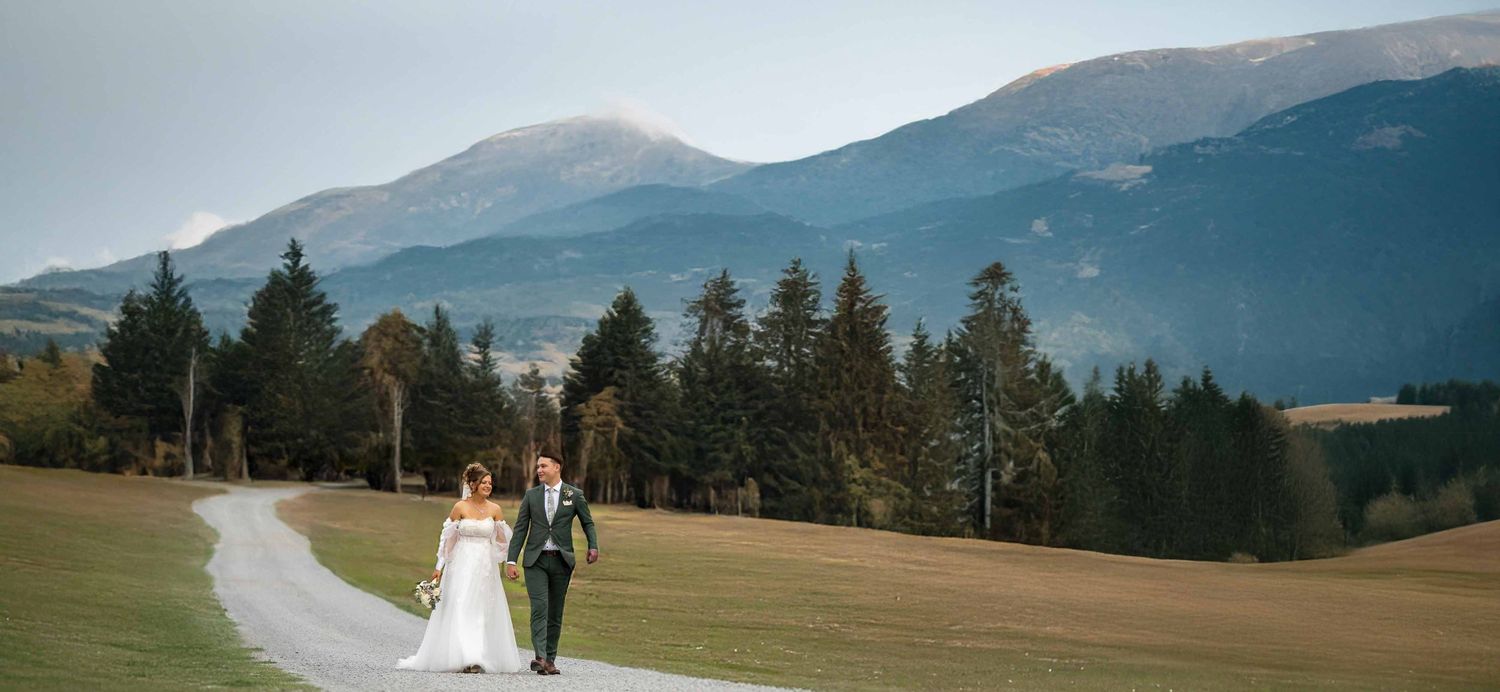 Bride and groom walk hand-in-hand along a scenic mountain path on their wedding day.