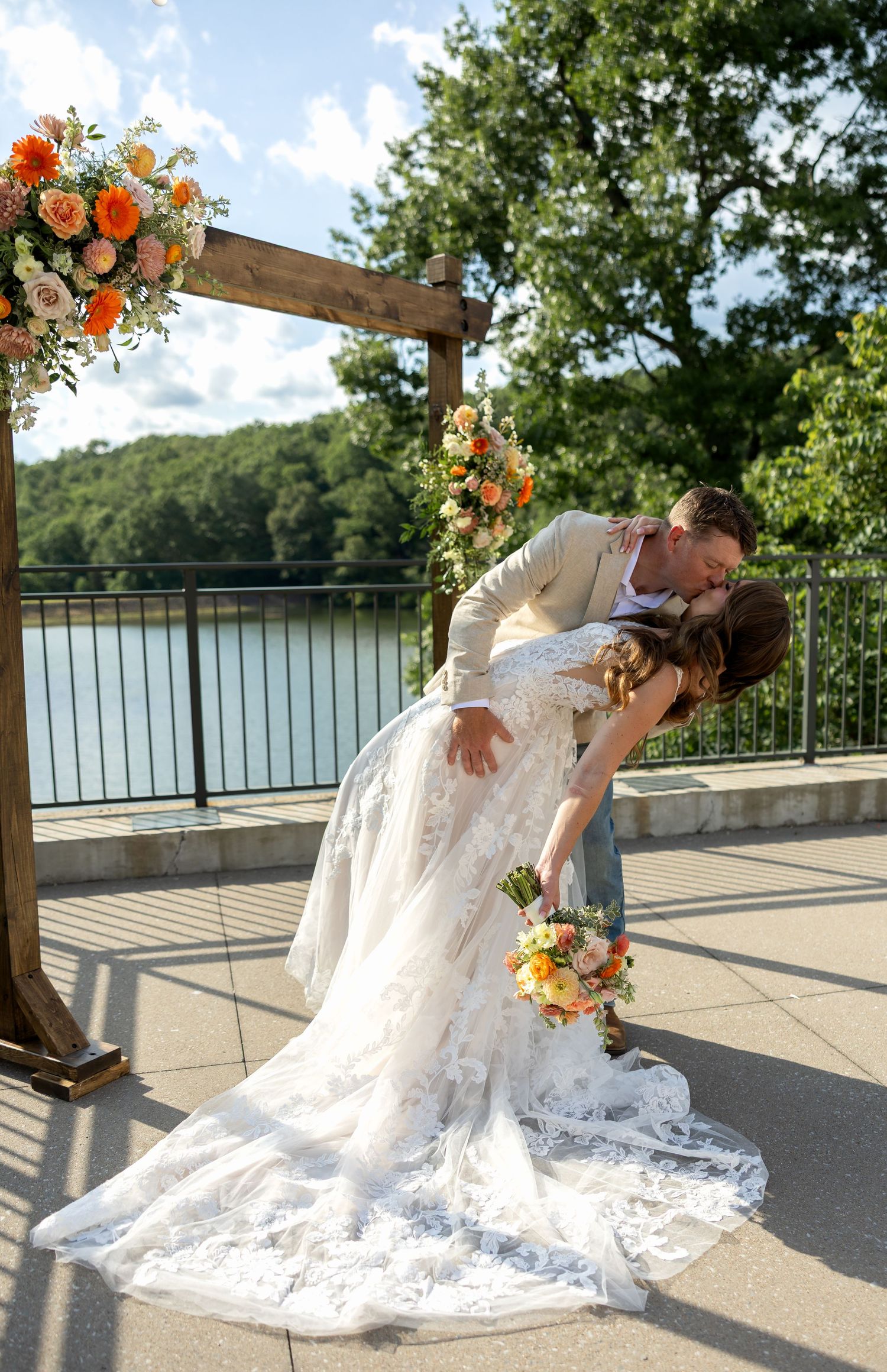 A romantic dip kiss under a wooden arch decorated with orange and white flowers by a lakeside.